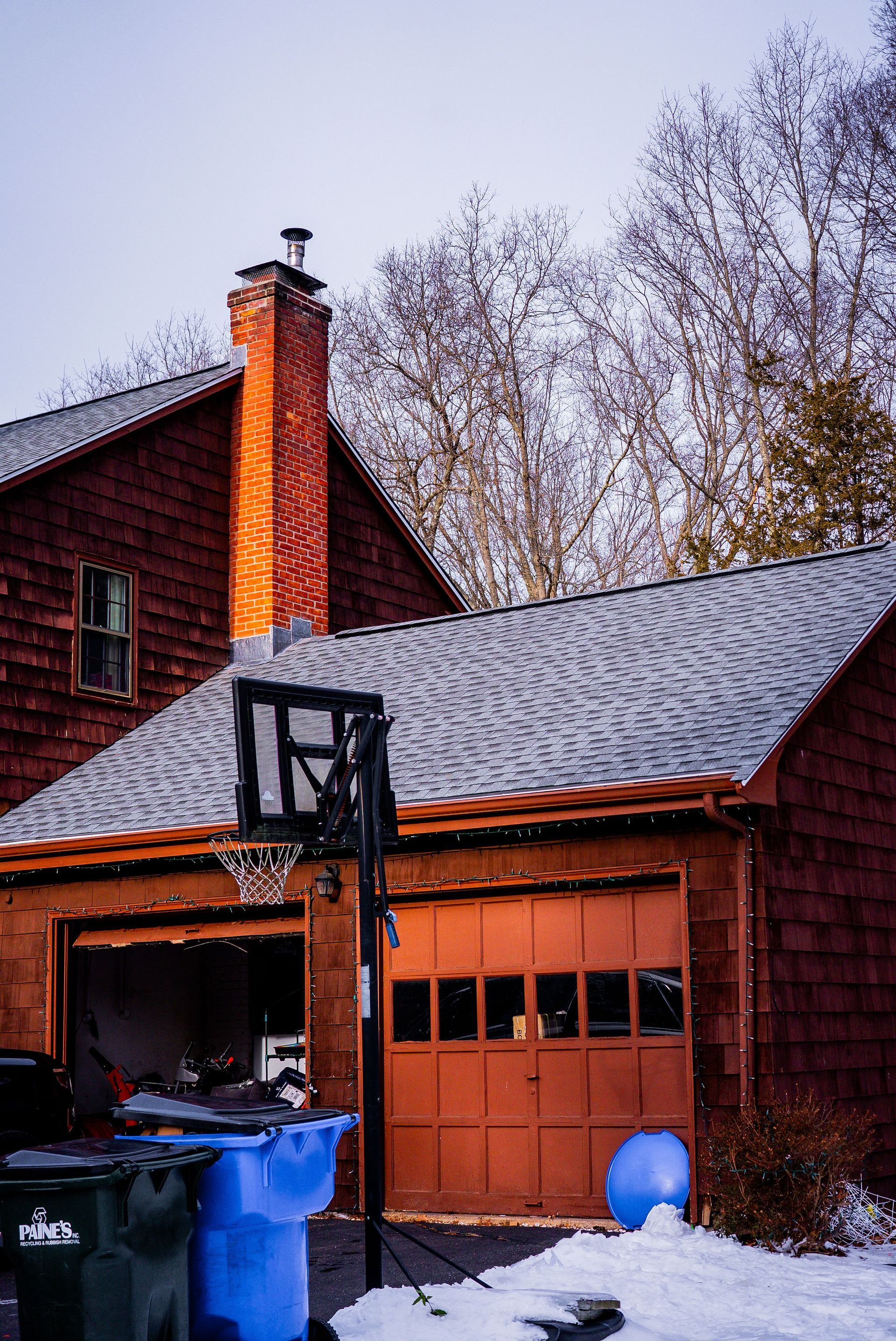 Red house with a garage, basketball hoop, and trash cans. Snow on the ground.