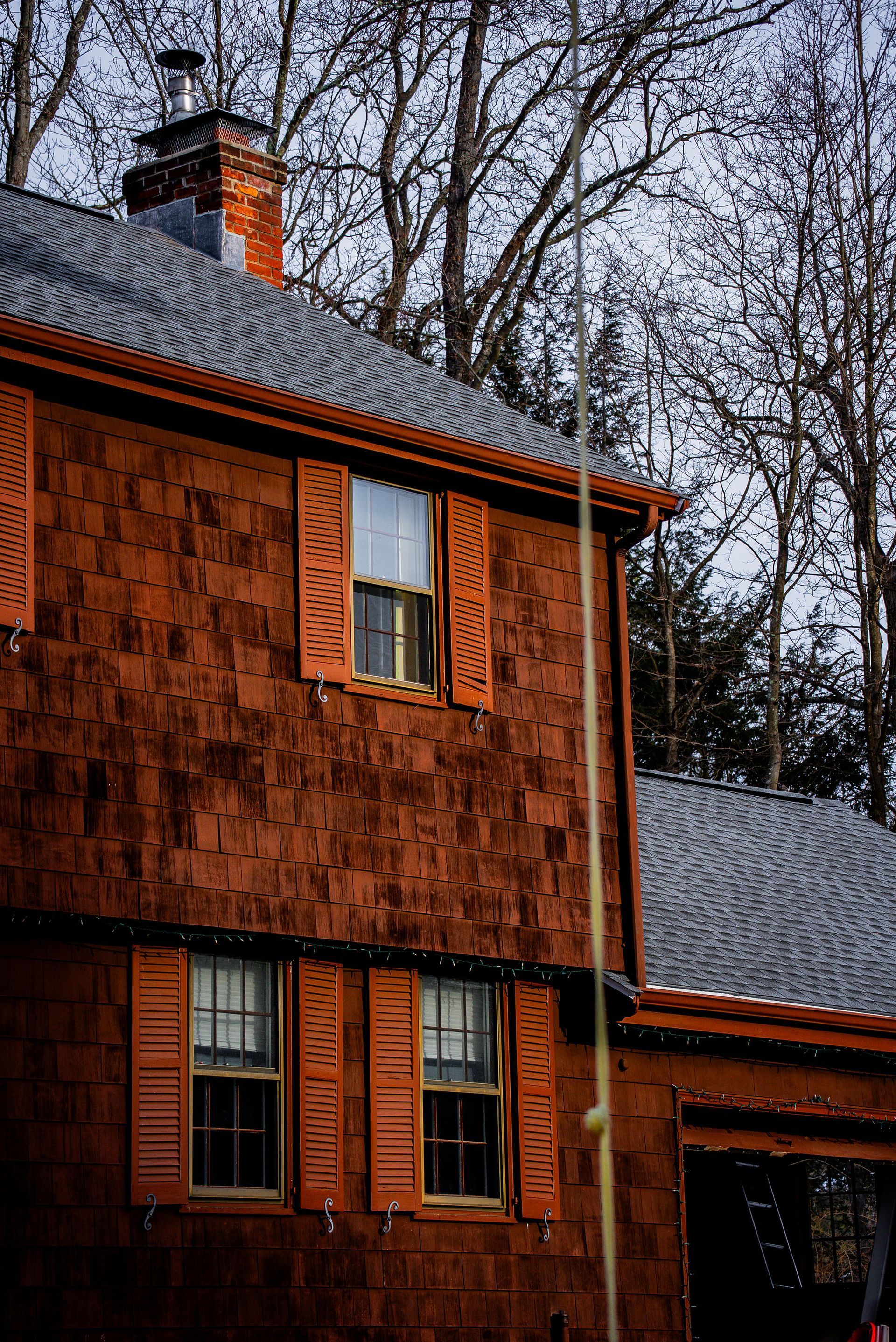 Red shingle house with open shutters; gray roof, brick chimney, and bare trees in the background.