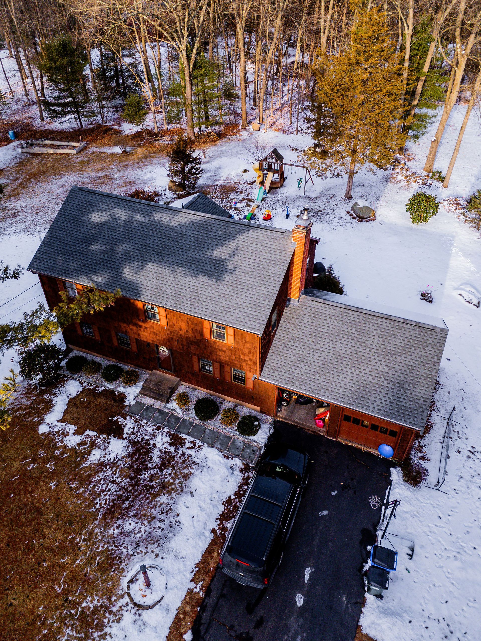 Aerial view of a two-story brick house with a dark driveway, snowy yard, and surrounding trees.