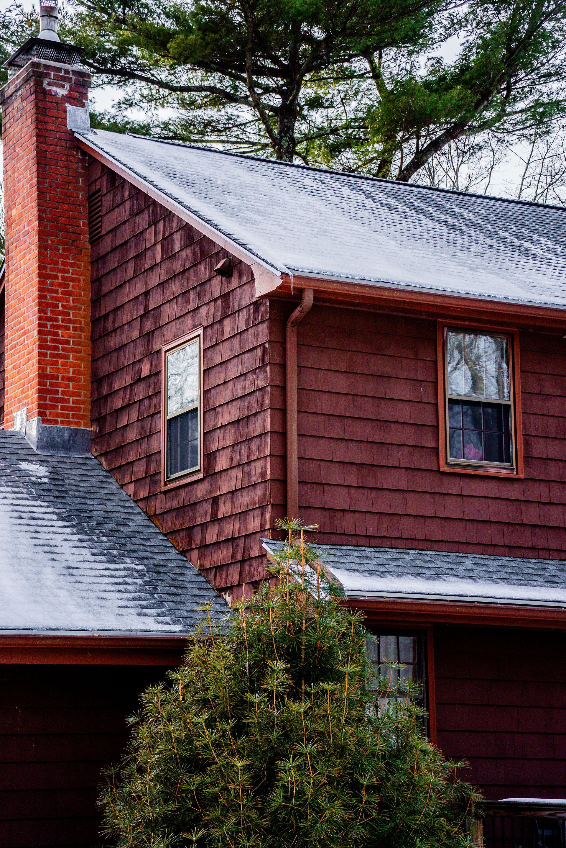 Red house with chimney and dark shingle roof, windows, and small evergreen tree in front.