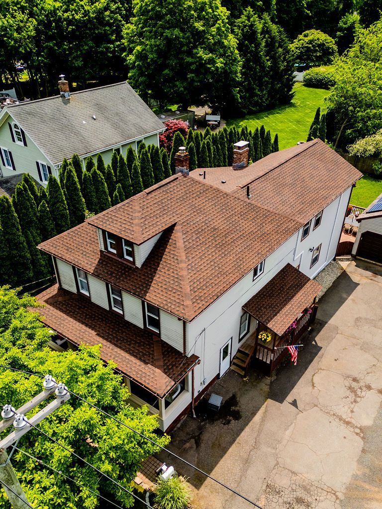 Bird's-eye view of a two-story white house with brown roof and porch, surrounded by trees and green lawn.
