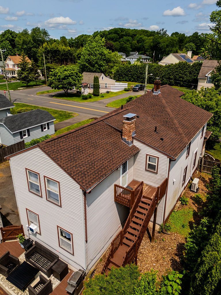 Exterior view of a two-story house with brown roof and wooden deck. Surrounded by trees and other houses.