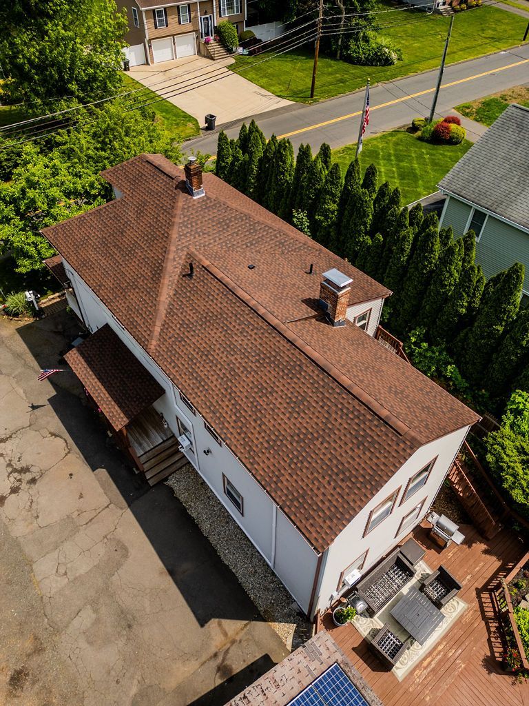 Aerial view of a white two-story house with a brown shingle roof, surrounded by greenery and parking.