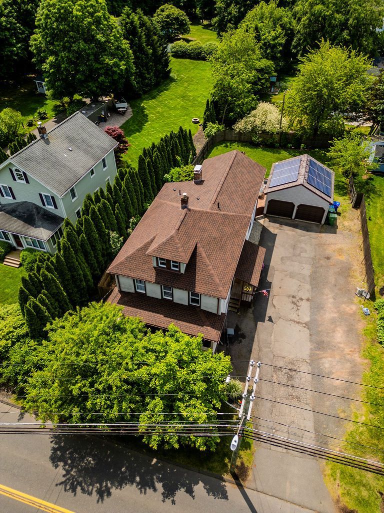 Aerial view of a house with a brown roof and a detached garage with solar panels. Green trees and lawn surround it.