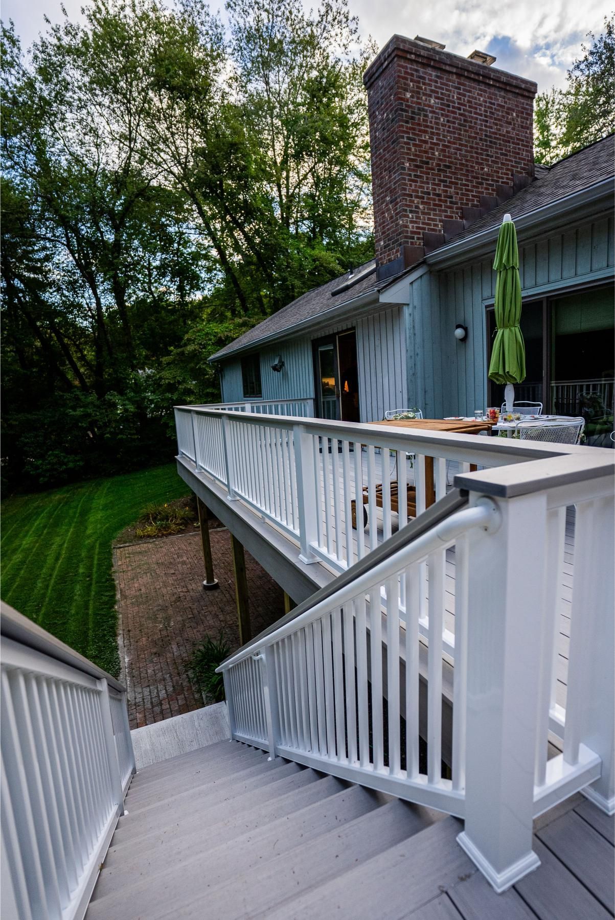 Wooden deck with stairs leading up to a house with a brick chimney and green umbrella.