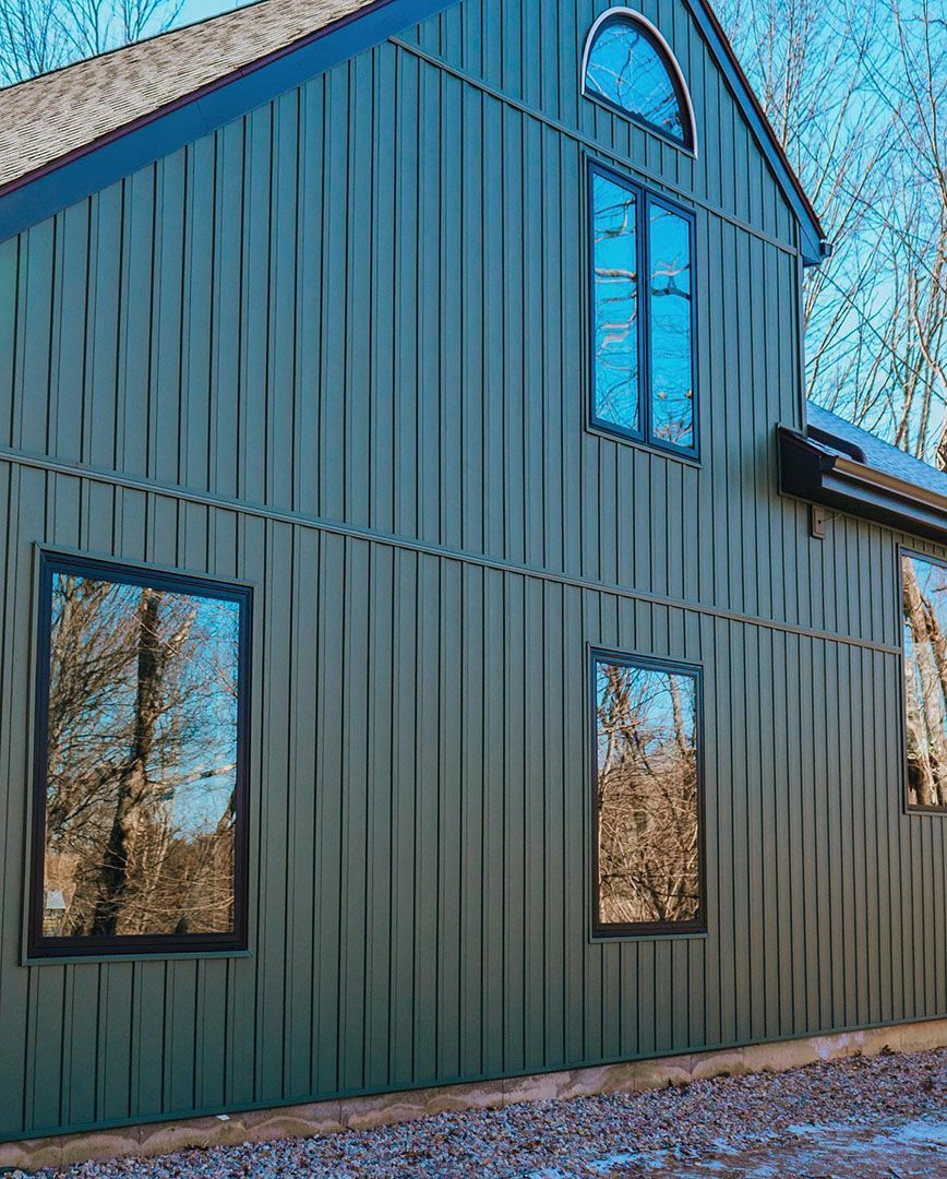 Dark green, two-story house with vertical siding and black-framed windows; trees reflected in the windows.