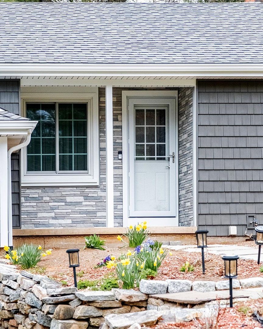 Exterior of a house with a door, window, gray siding, and a stone retaining wall.