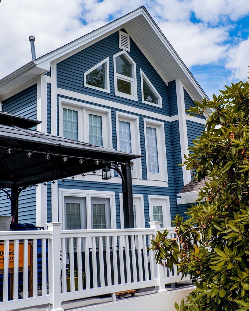 Blue two-story house with white trim. Deck and pergola in foreground, sunny sky.