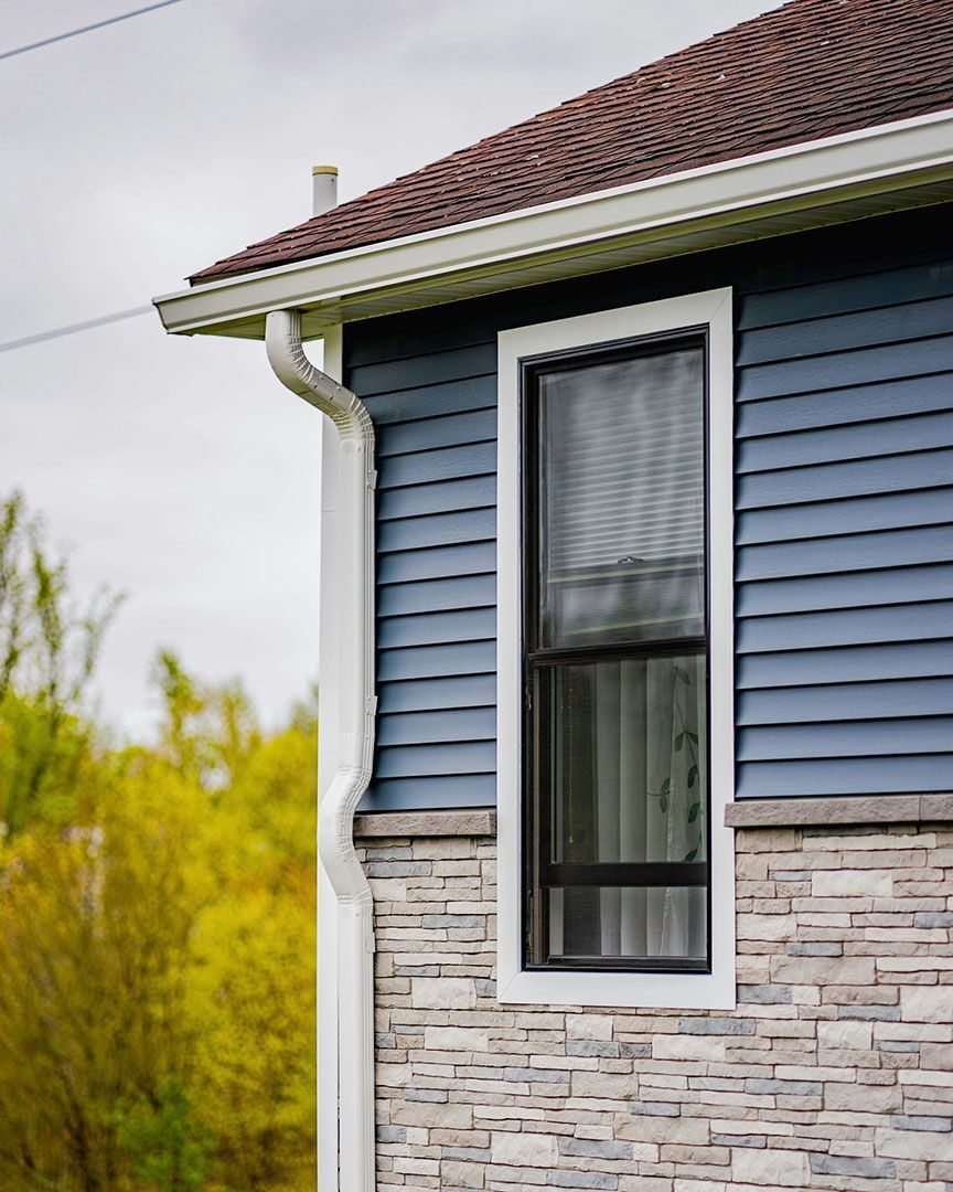 Side view of a house with blue siding, stone facade, and a tall window. White trim and gutters.
