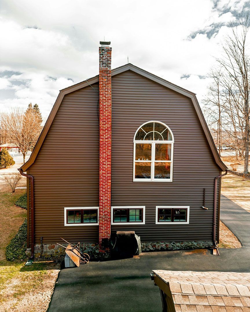Brown barn-shaped house with a red brick chimney, arched window, and rectangular windows.