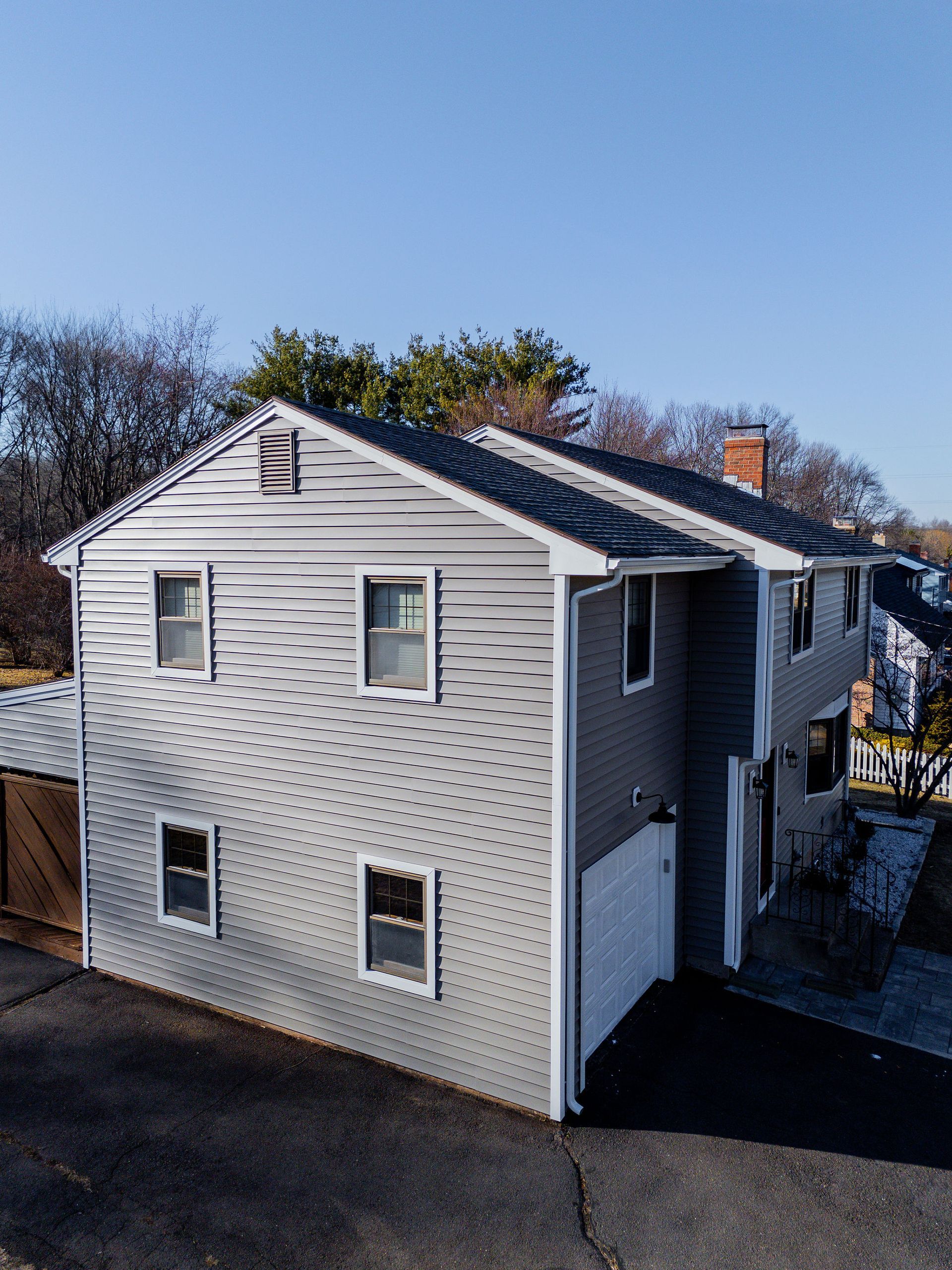 Two-story gray house with white trim, four windows visible, garage, and chimney, under a clear blue sky.