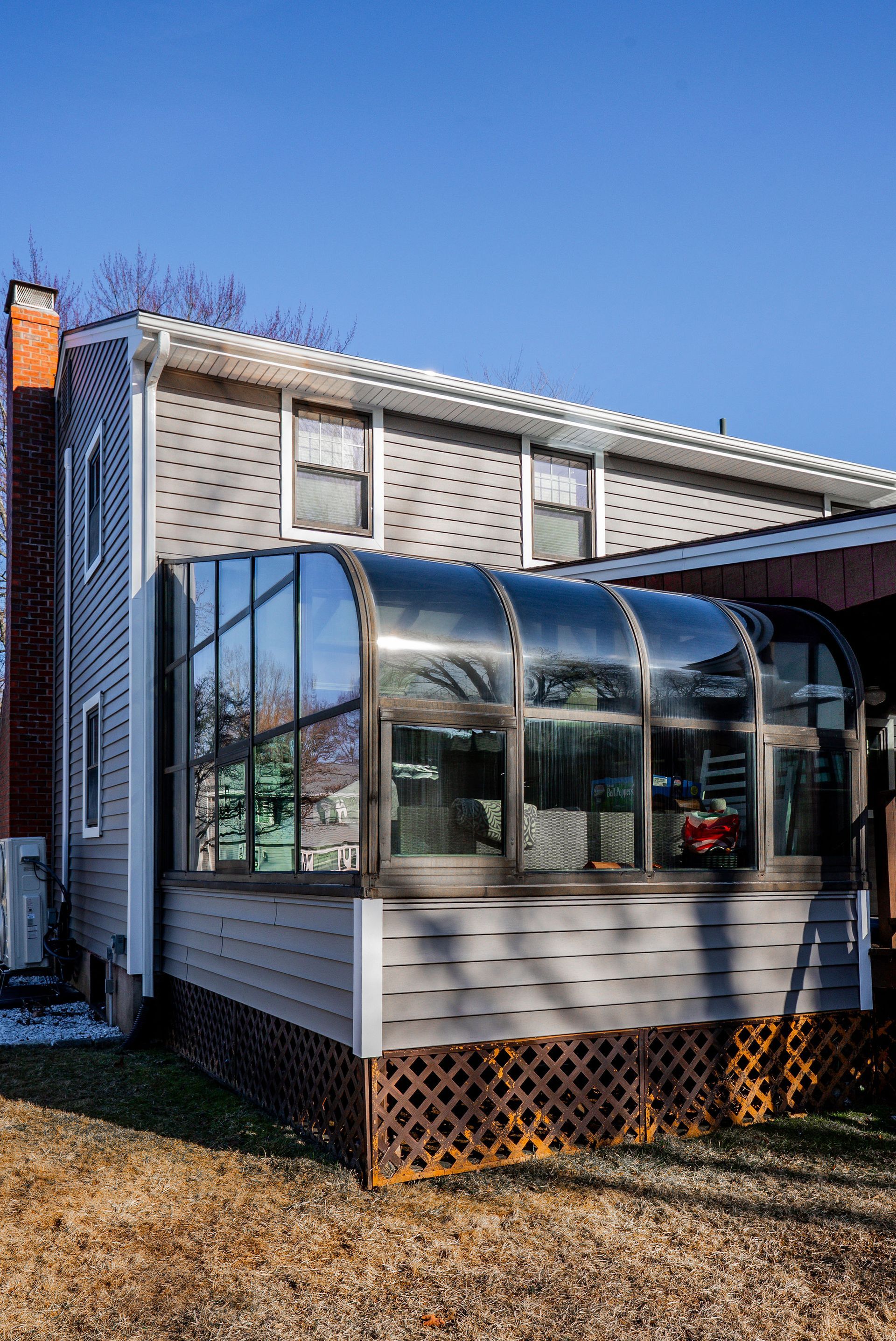 Sunroom addition on a gray house with a curved glass roof and latticework base; sunny day.