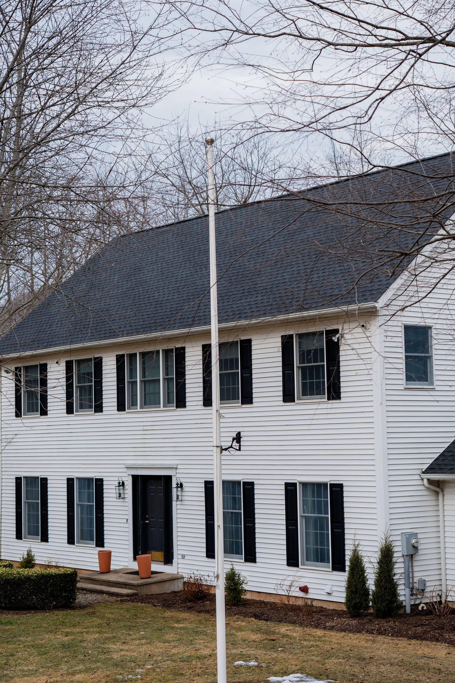 White two-story house with black shutters, tall antenna in front, overcast sky.