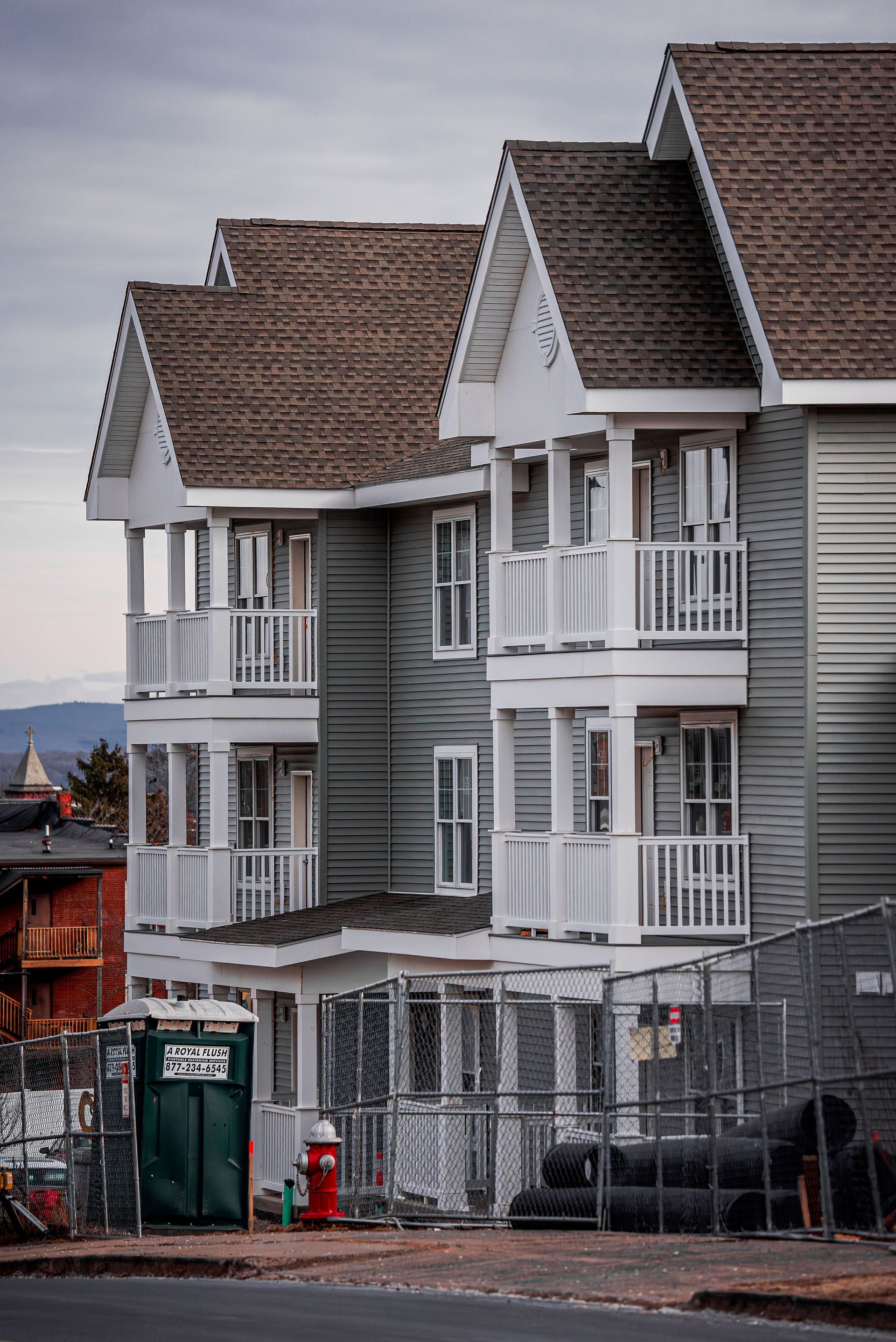 Three-story light blue building with white balconies and brown roof, next to a portable toilet and a fence.