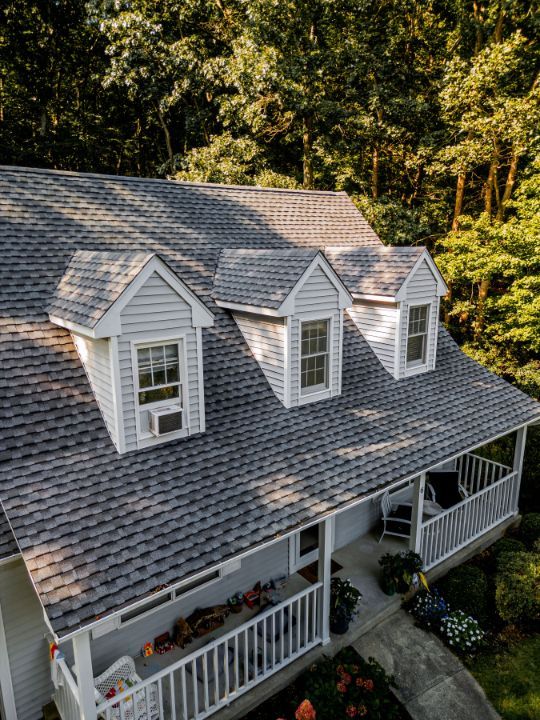 House with gray shingled roof, white siding, three dormers, and a porch, set amongst trees.