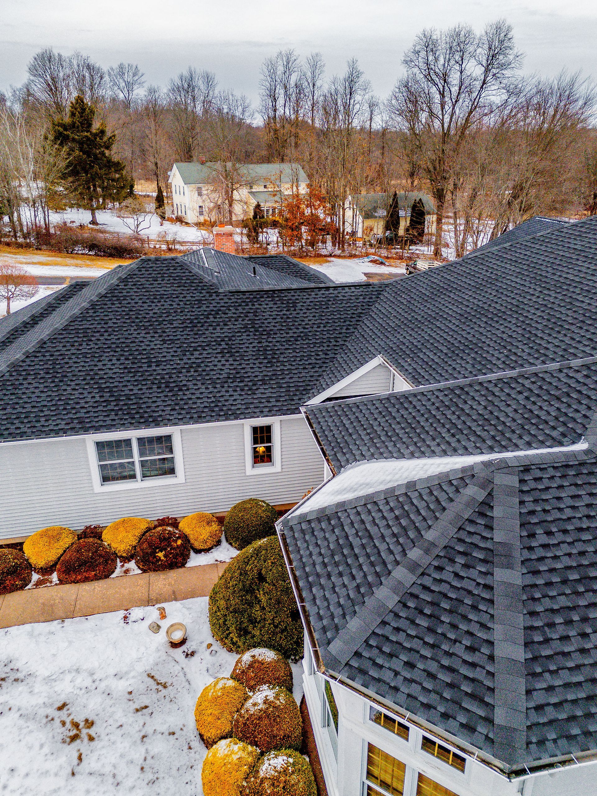 Dark gray shingled roofs of a house in winter, overlooking a snow-covered yard and distant houses.