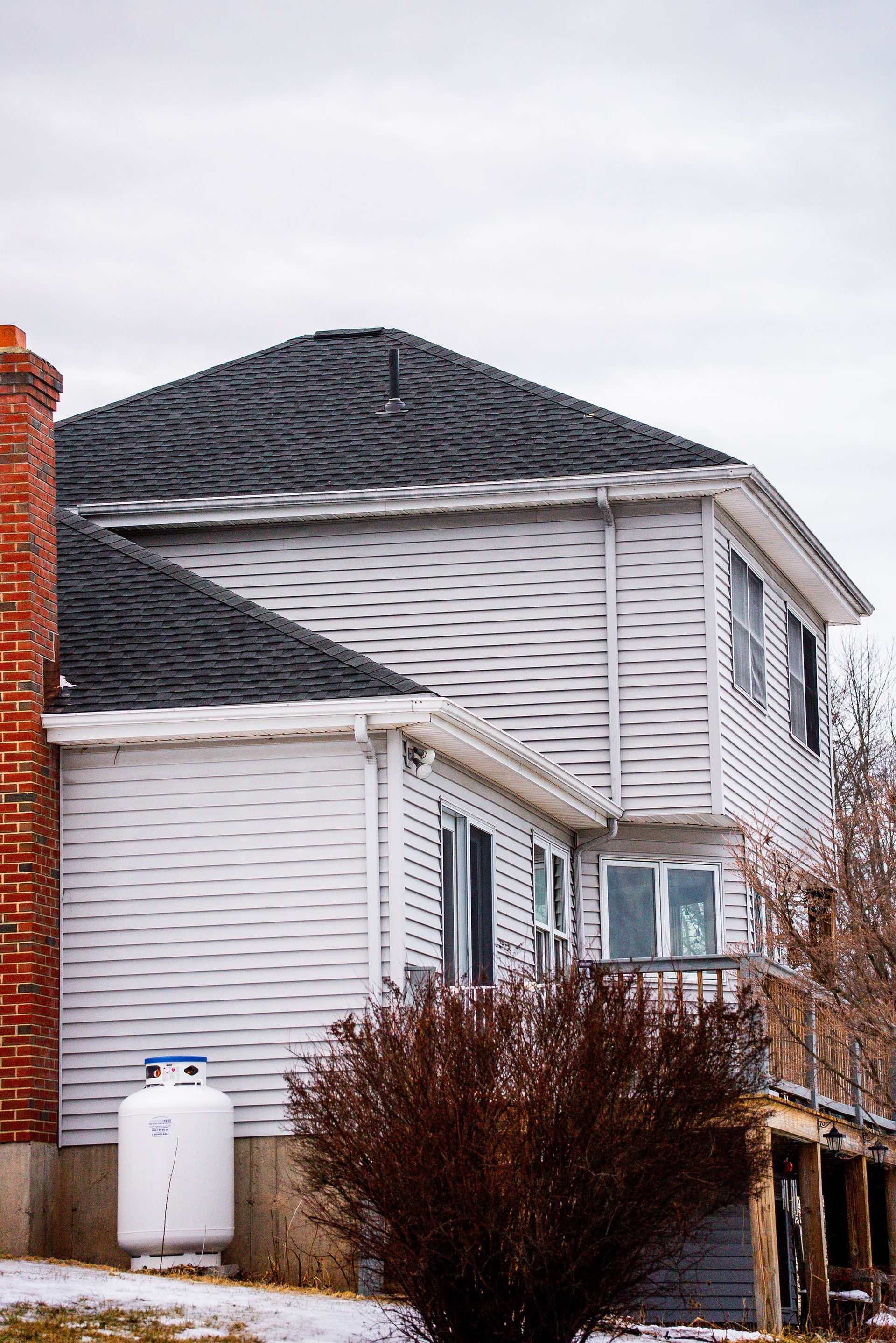 White two-story house with black roof, brick chimney, propane tank, and brown bush in front. Cloudy sky.