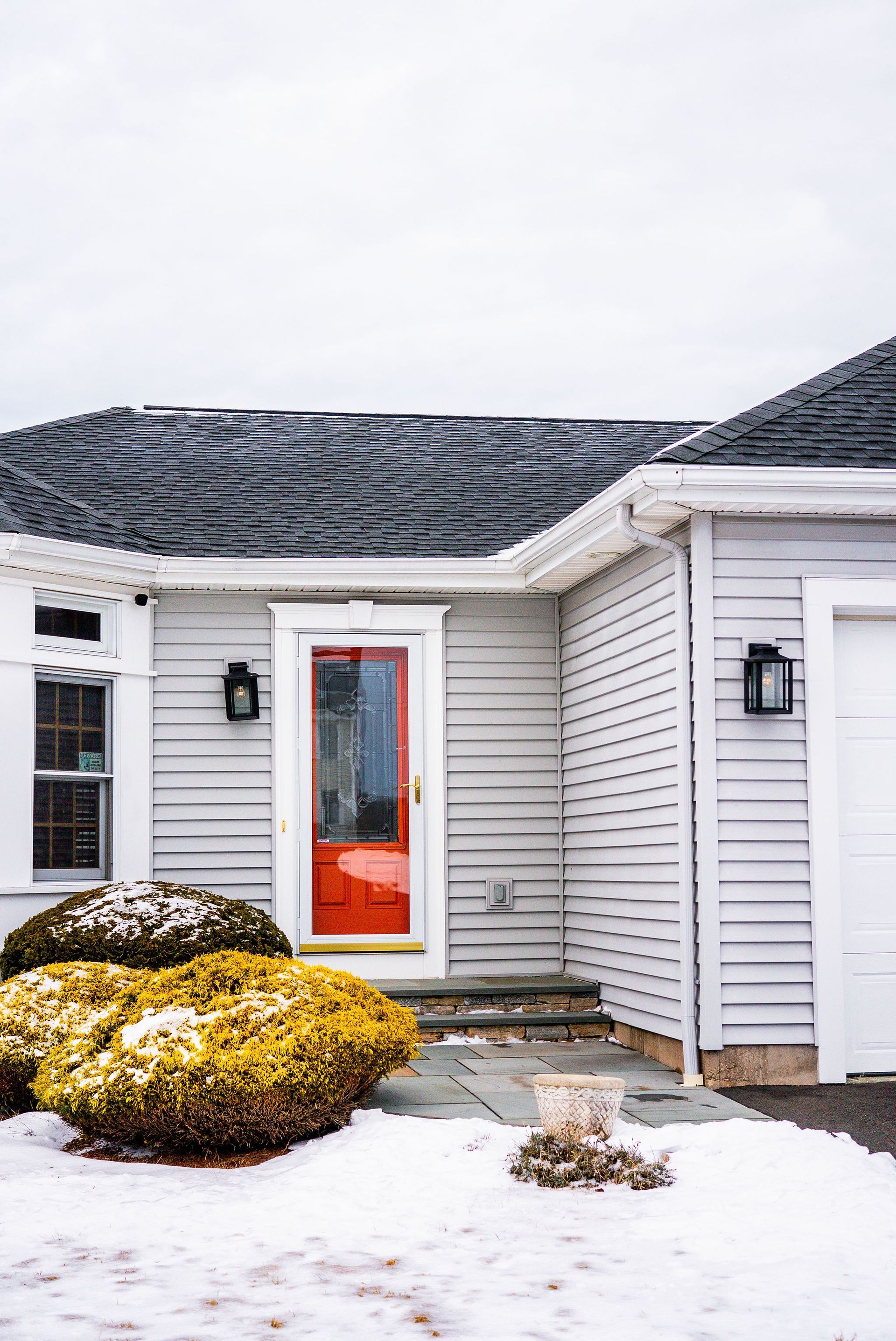 Front of a house with a red door, white siding, black roof, and snow-covered landscaping.