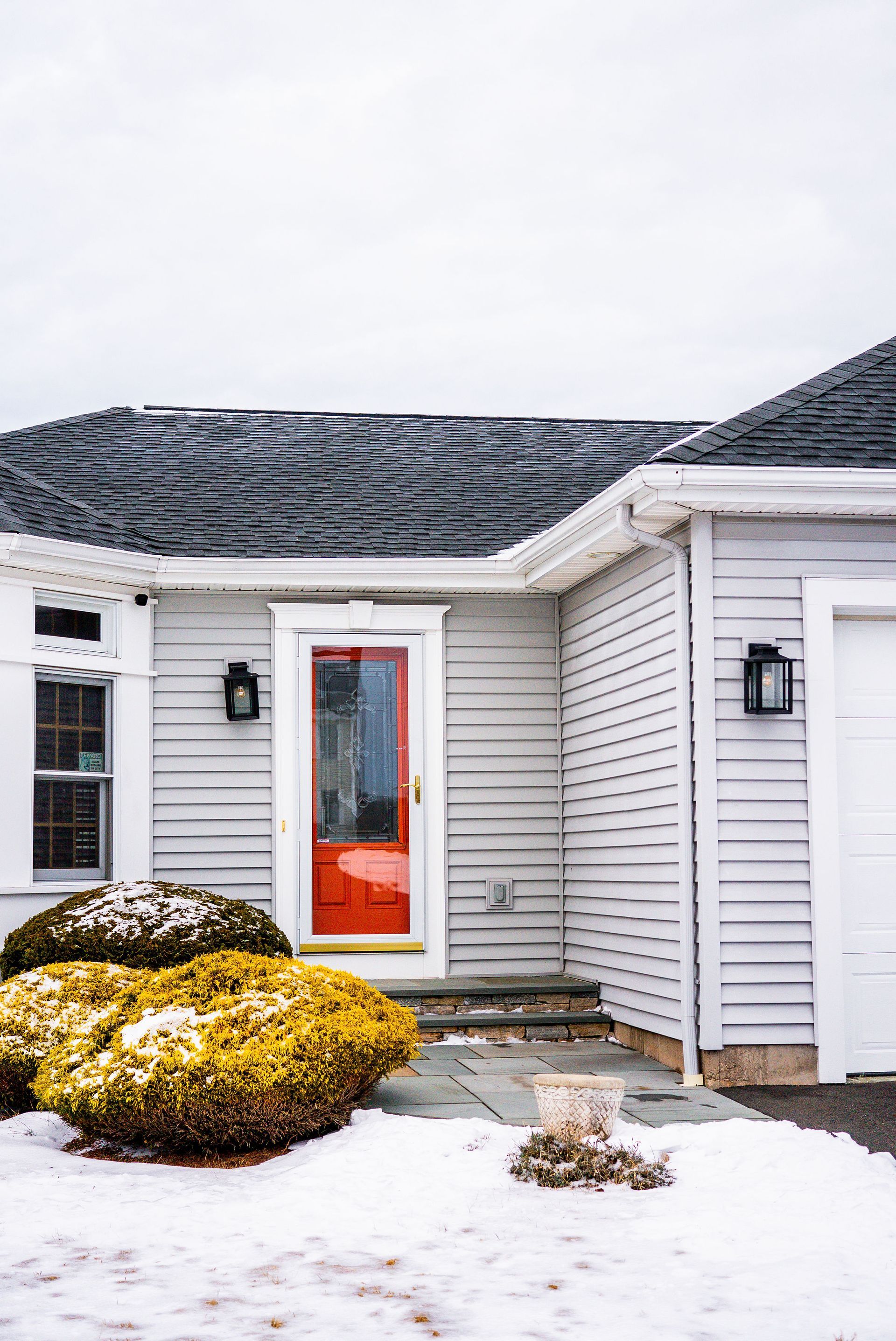 White house with a red front door, surrounded by snowy ground and shrubs.