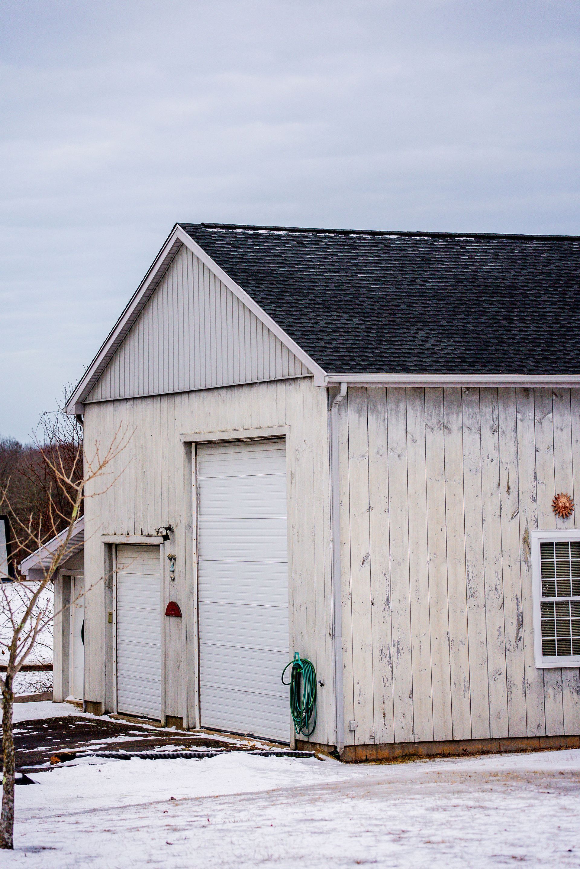 White weathered shed with dark roof and snow. Garage door, side door, and window.