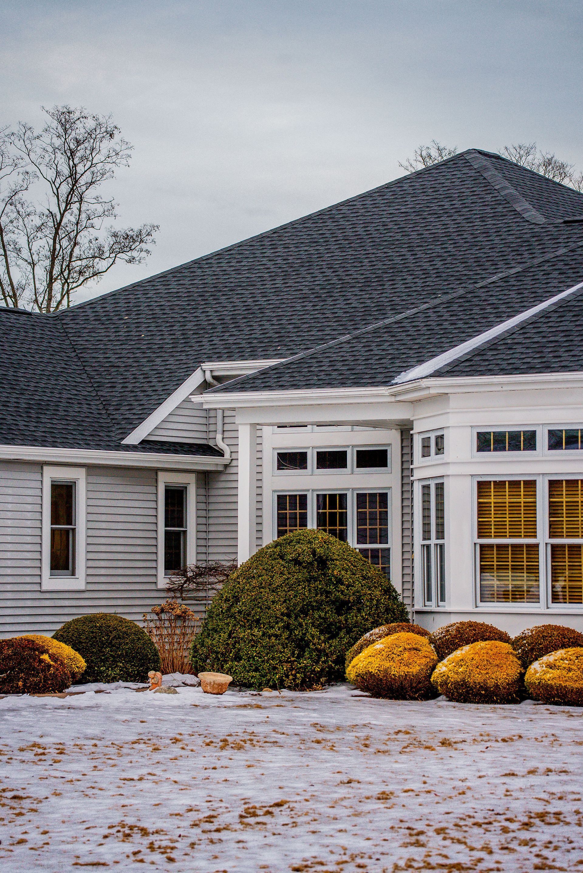 Gray house with dark roof and white trim. Yellow and green bushes in front on a snowy lawn.