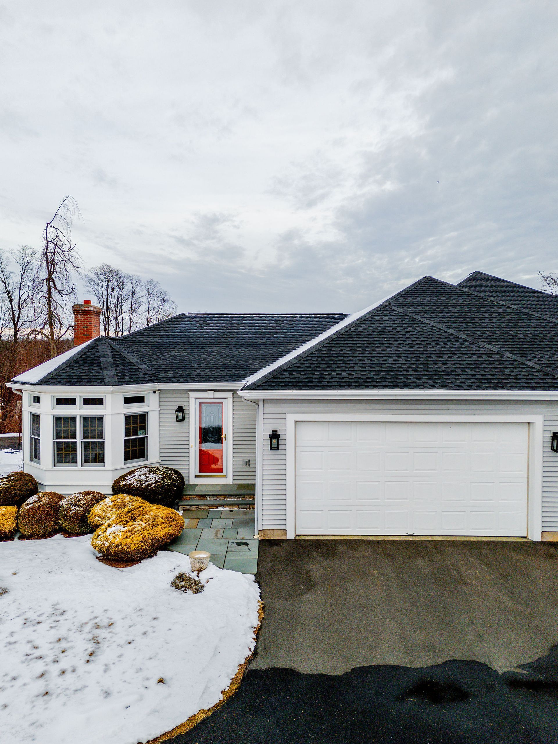 White house with black roof, red door, garage, snowy yard, and cloudy sky.