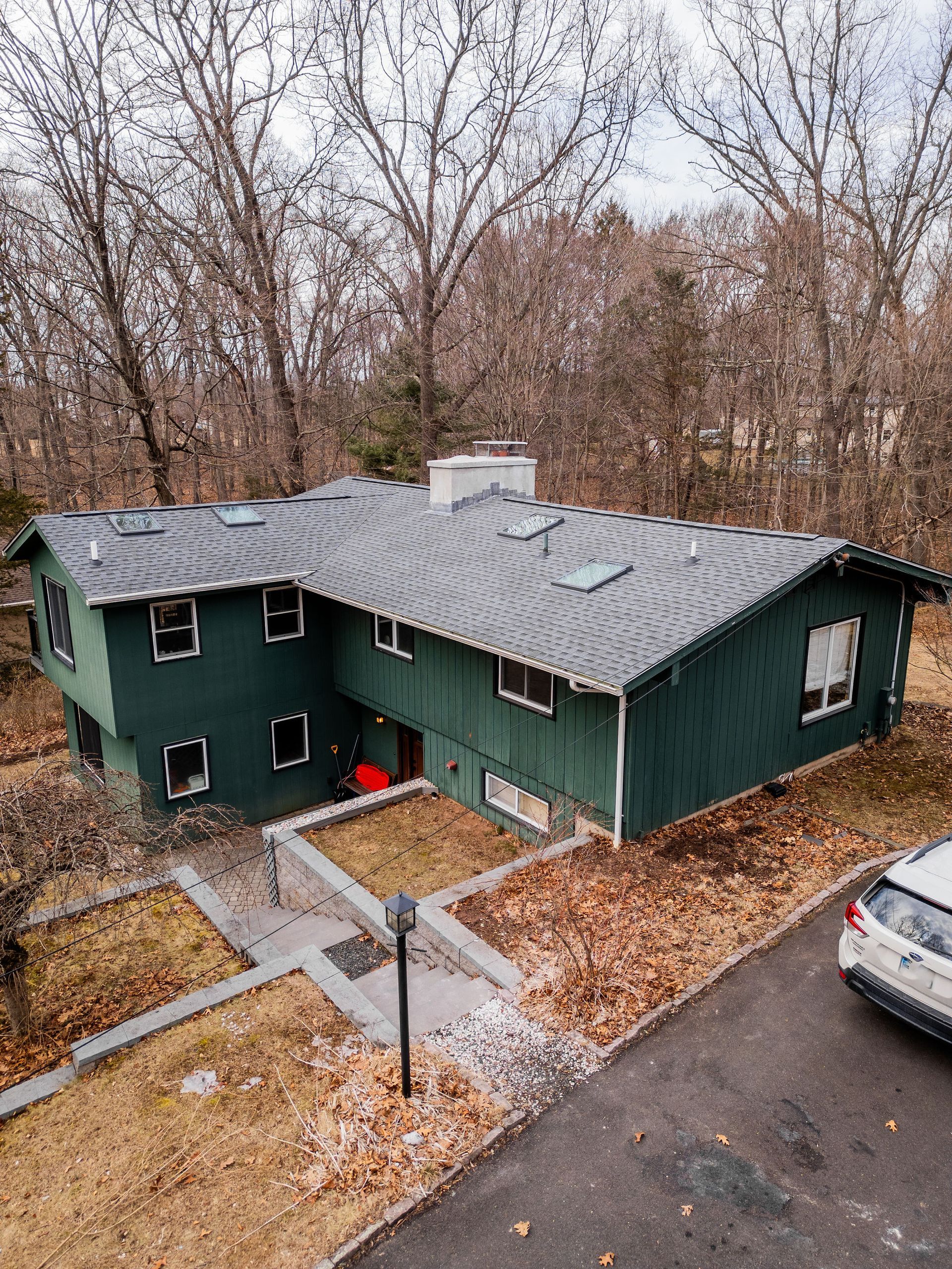 Green house with dark roof and surrounding trees; parked car in driveway.