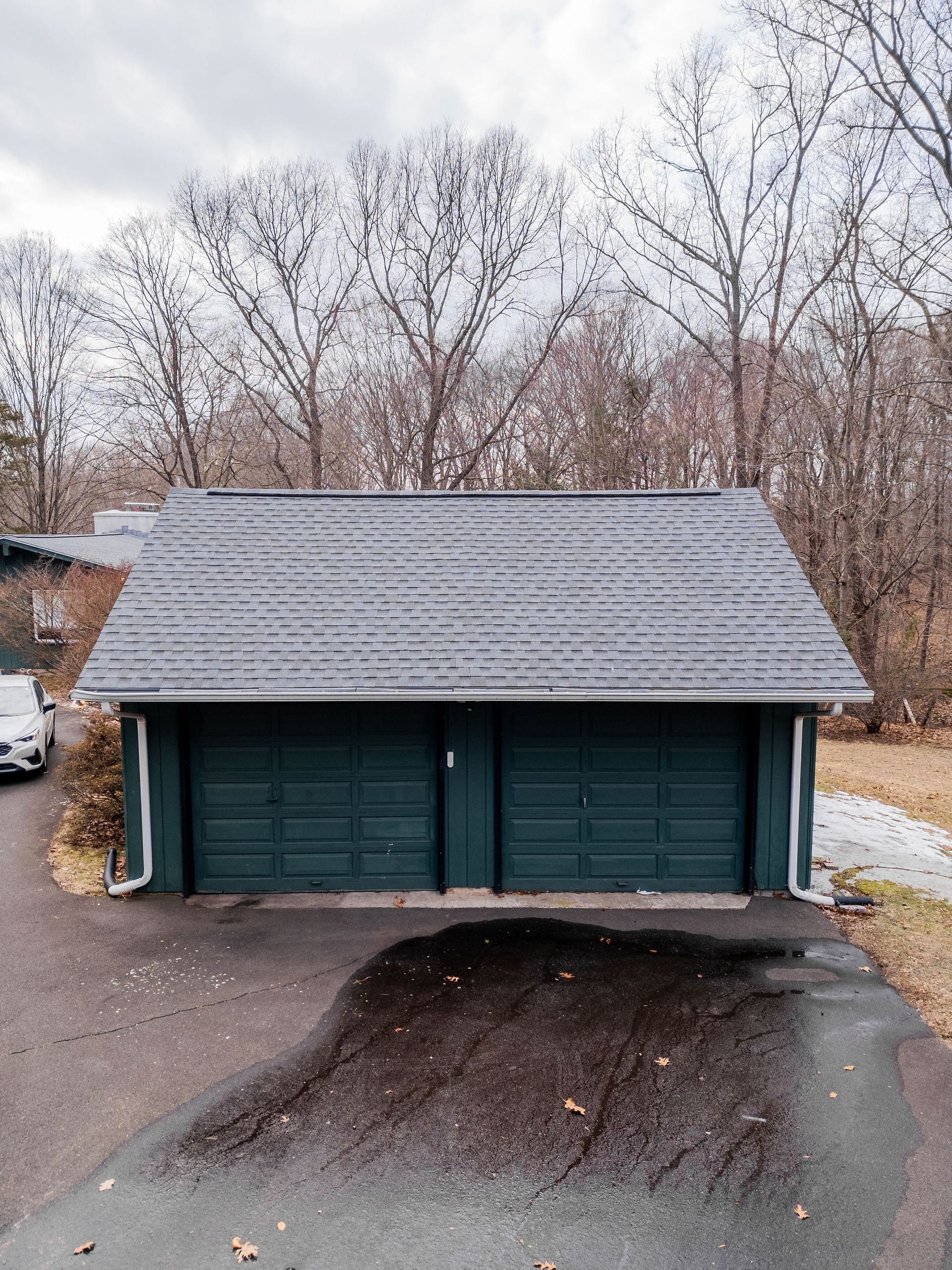 Green double garage with dark roof, located on a paved driveway with bare trees in the background.