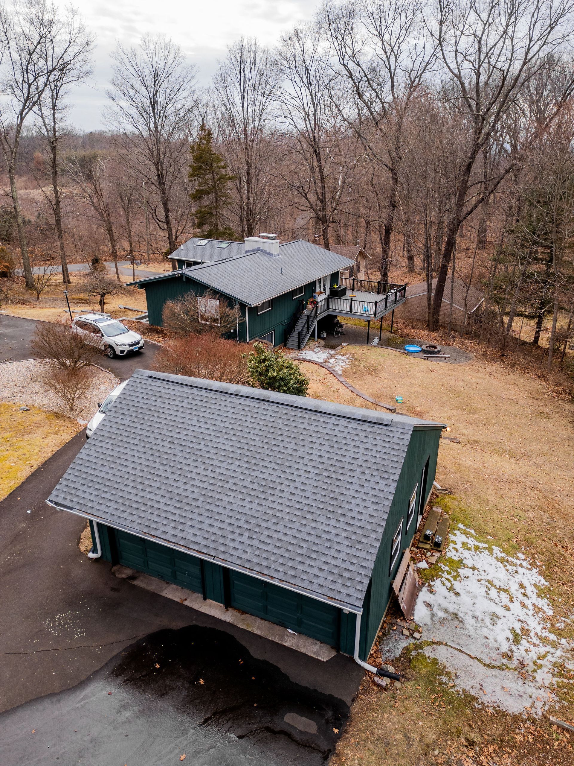 Green house and detached garage on a driveway, set among bare trees in winter.
