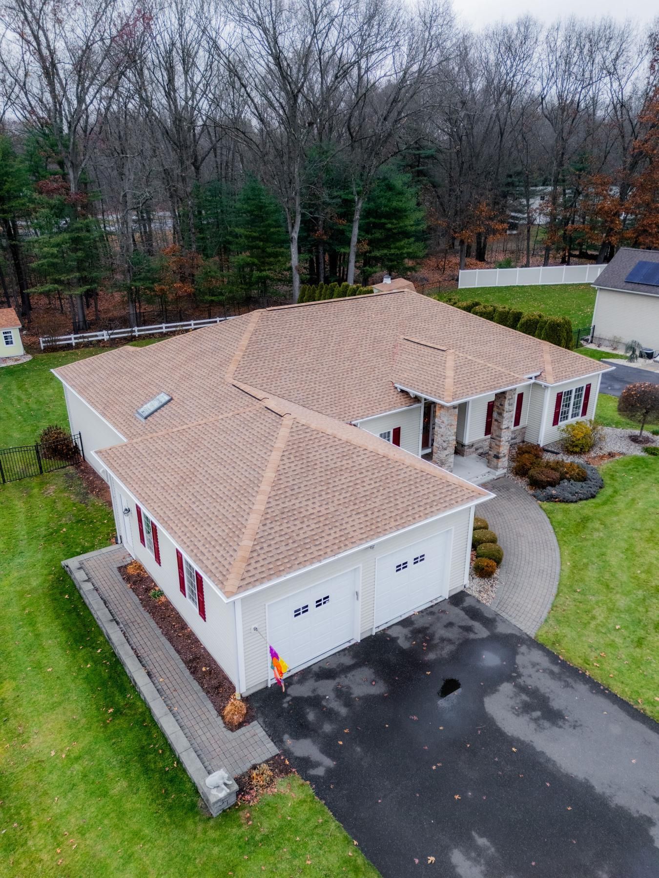 Aerial view of a beige house with a brown roof and attached garage, surrounded by green grass and trees.