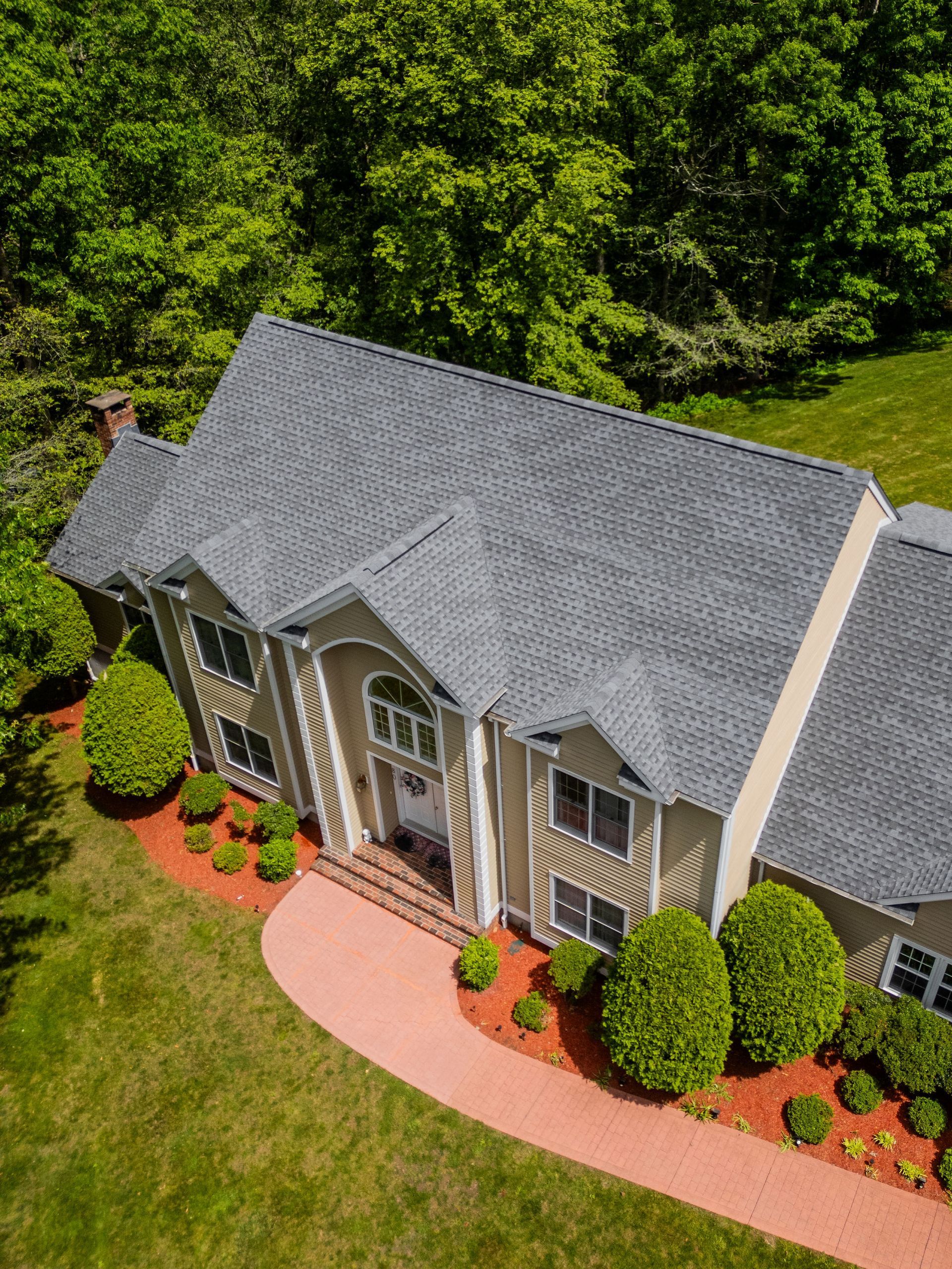 Aerial view of a beige house with a gray roof and a red brick pathway surrounded by green bushes and grass.