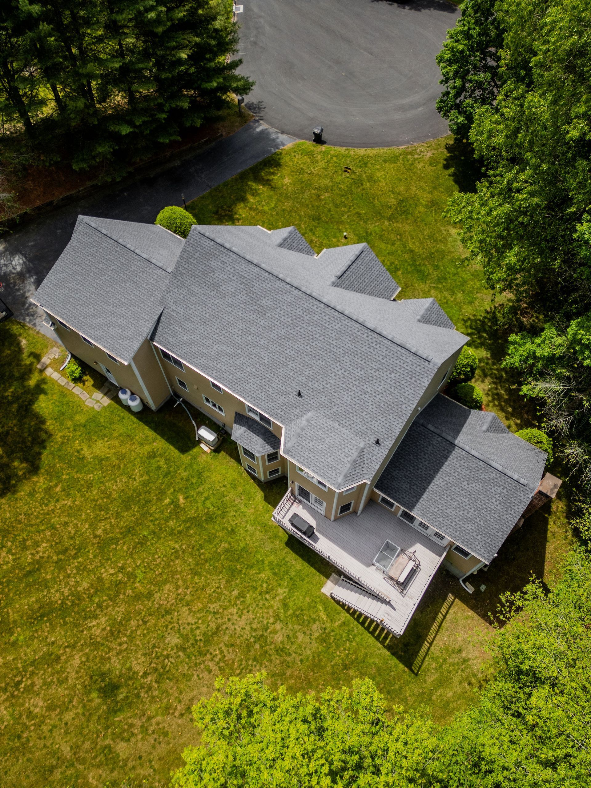 Aerial view of a gray-roofed house with a deck on a green lawn surrounded by trees and a driveway.
