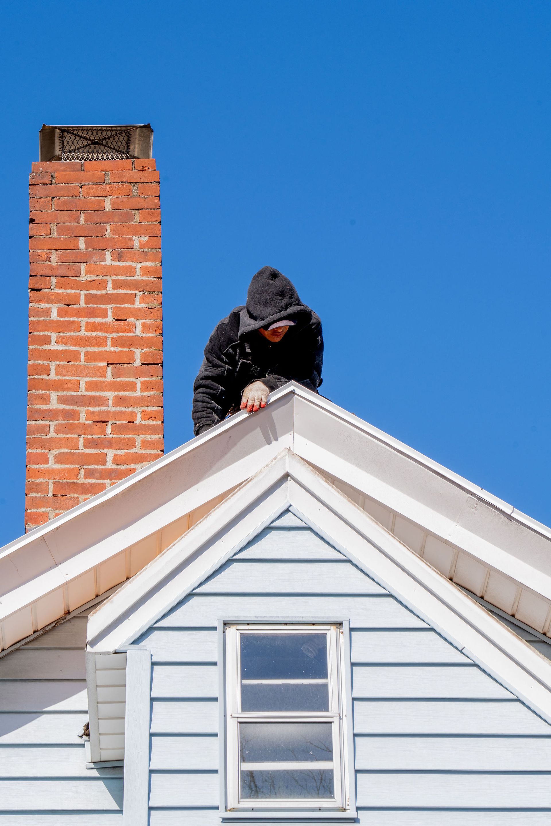Person in a hooded jacket on a roof, near a brick chimney, against a blue sky.