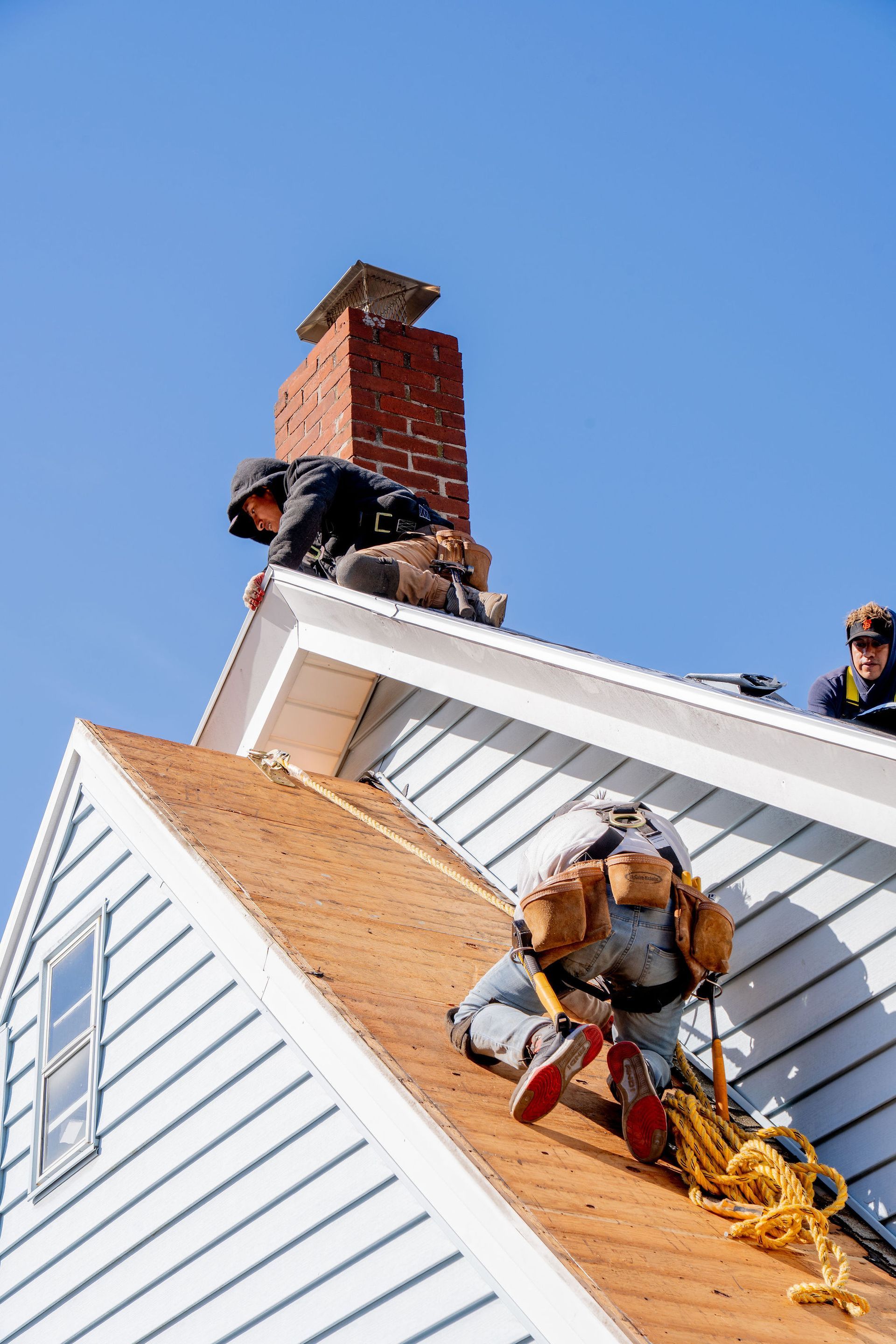 Roofers work on a house, one climbing, others near a chimney, against a blue sky.