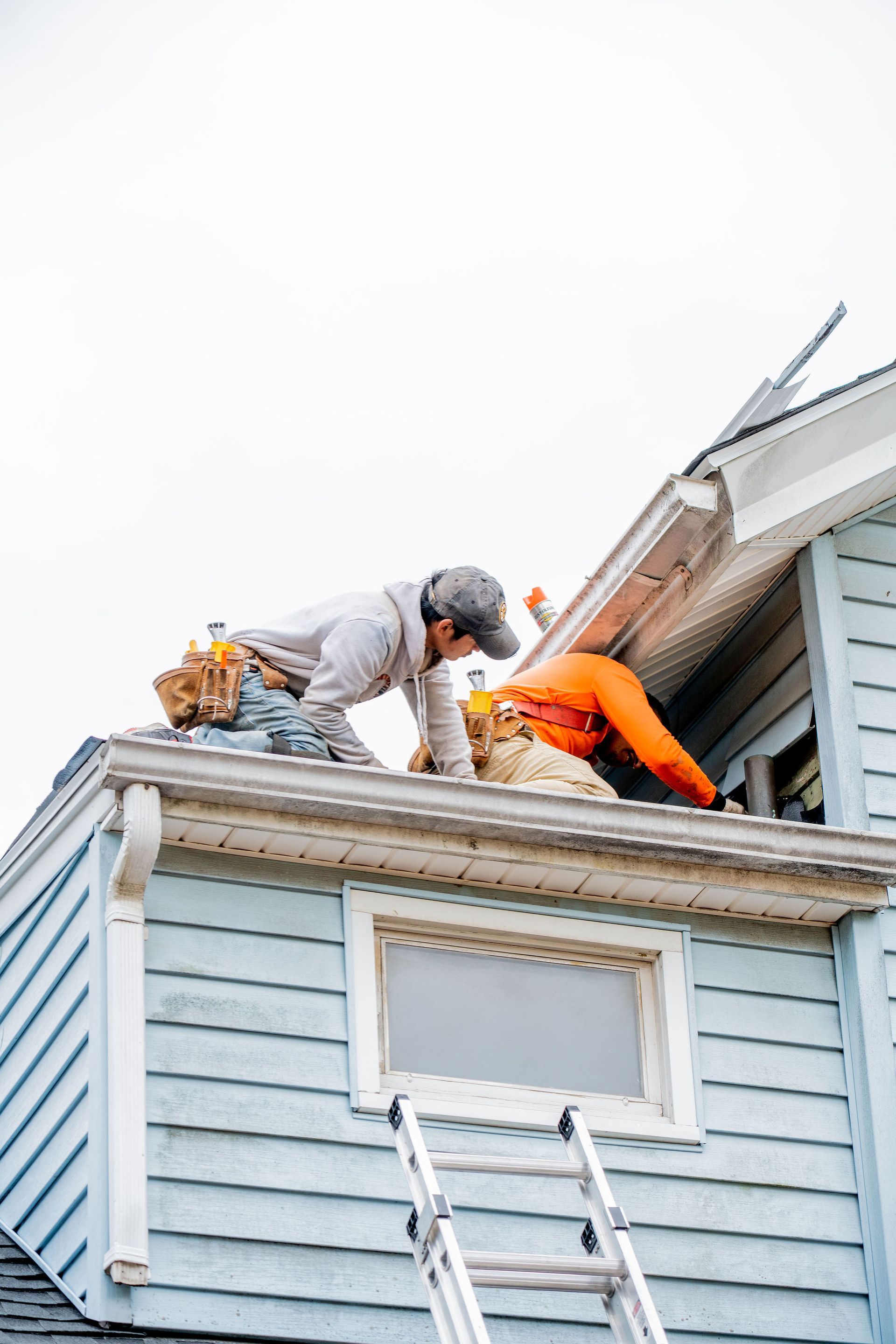Two people working on a roof, one in an orange shirt and the other in a gray hoodie, blue house, ladder.