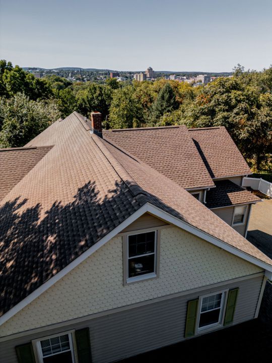Overhead view of a house with a brown shingle roof, green shutters, and a cityscape in the background.