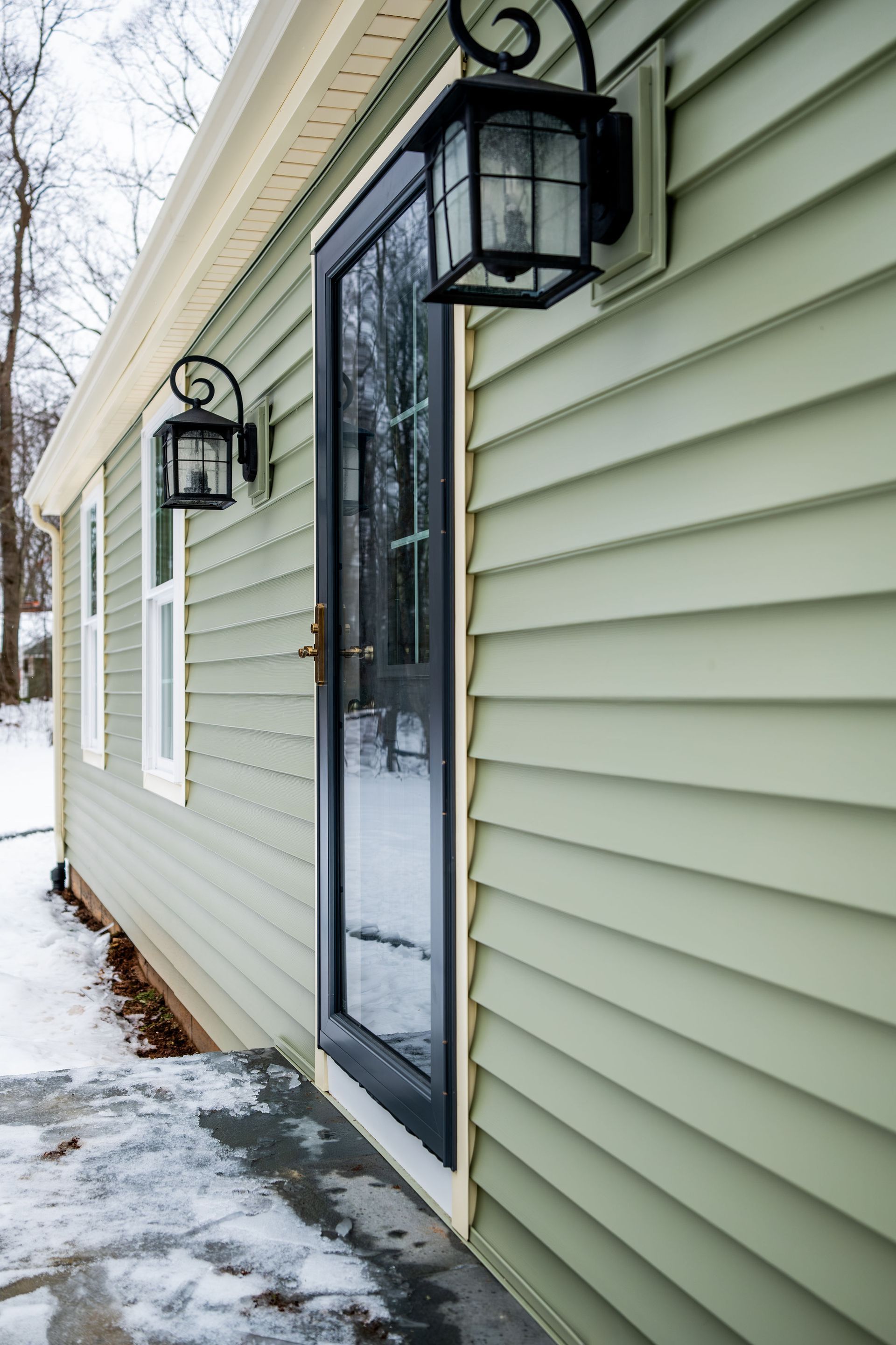 Green-sided house with black door and two sconce lights; snowy ground in the foreground.