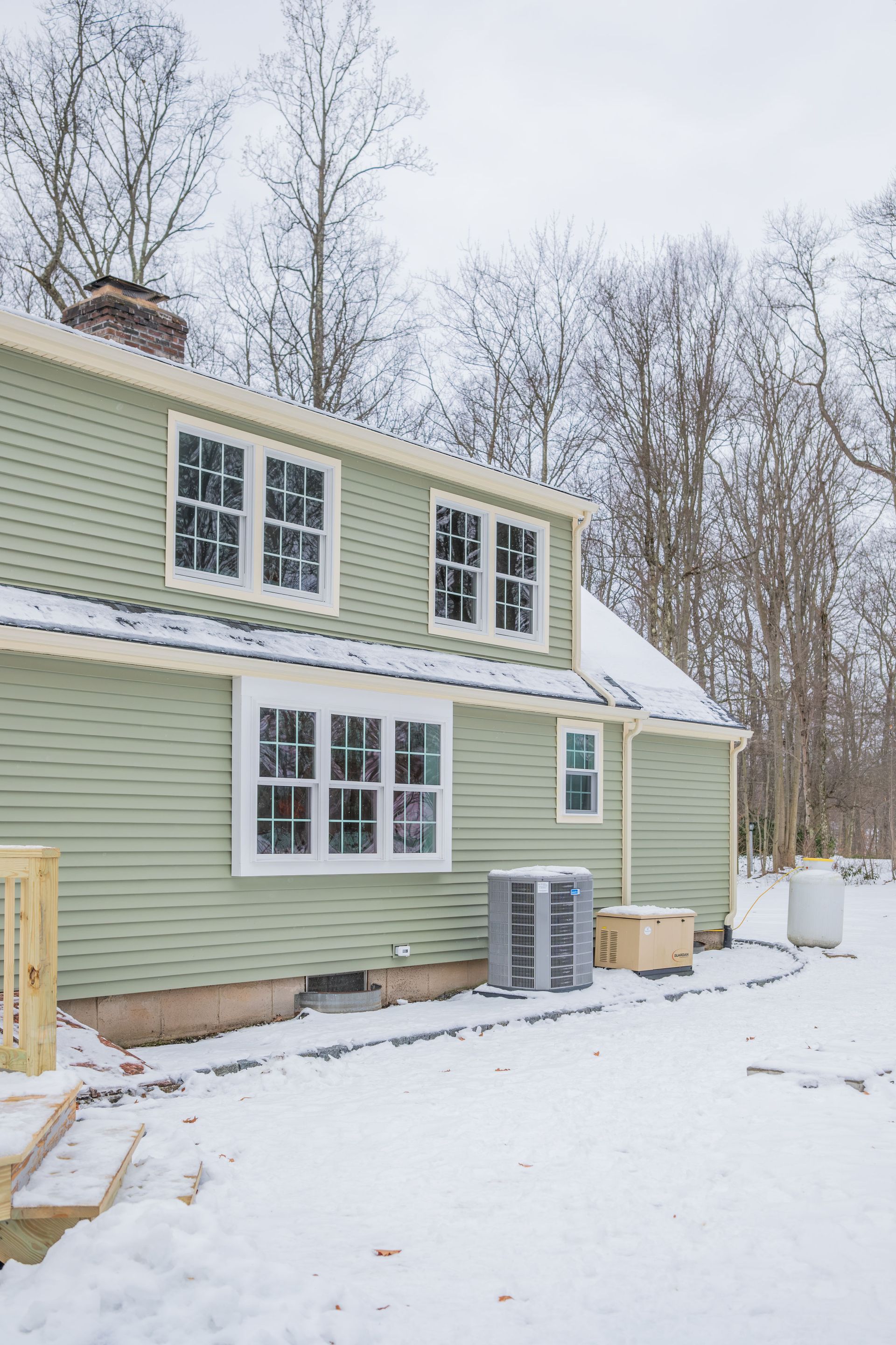 Two-story green house with white-framed windows, surrounded by snow-covered ground and trees.