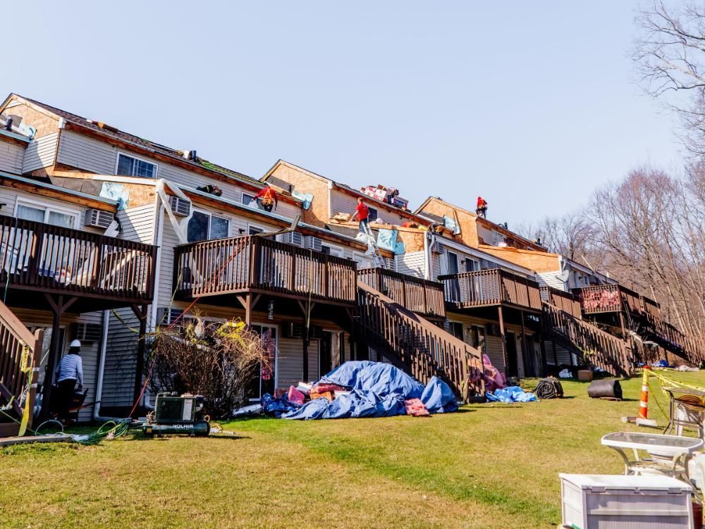 Row of townhomes with workers on roofs; repairing shingles. Blue tarp on the lawn below.