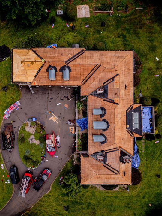 Overhead view of a house with fire trucks in the driveway, debris on lawn and roof partially covered by tarps.