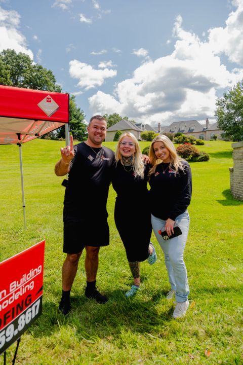 Three people smiling in front of a tent and grassy landscape. The person on the left is giving a peace sign.