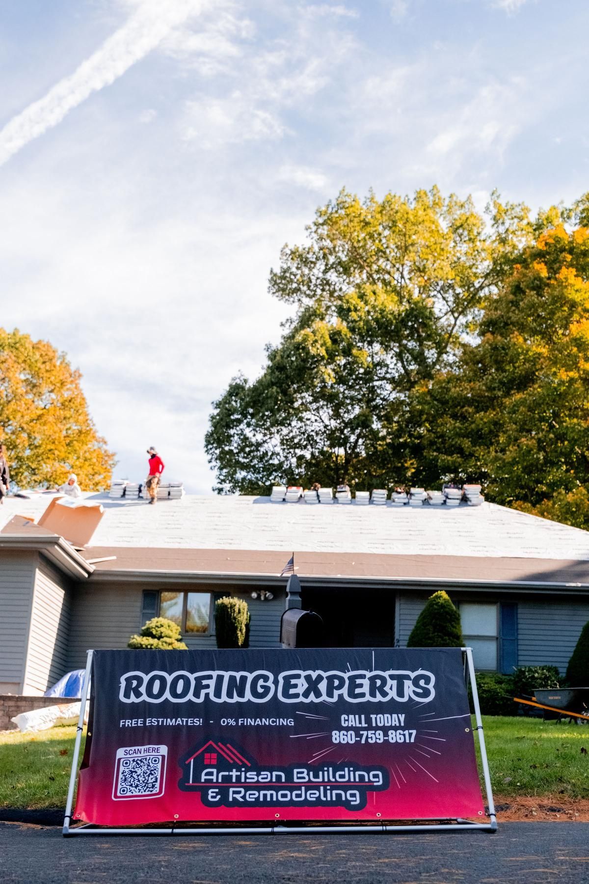 A roofing company sign in front of a house. A worker is visible on the roof. Bright sunny day.