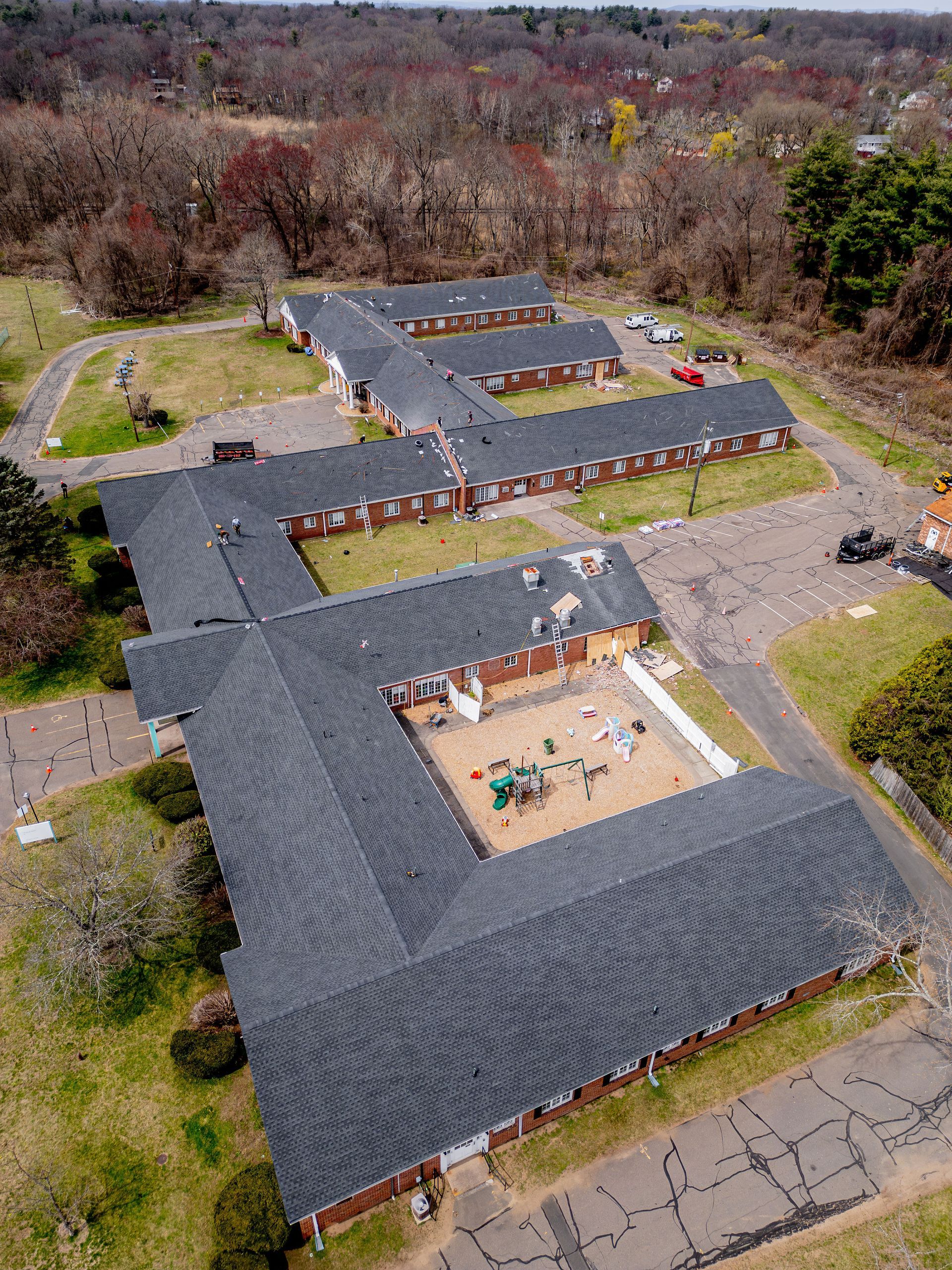 Aerial view of a long, brick building with a dark roof in a wooded area. Playground in the center.