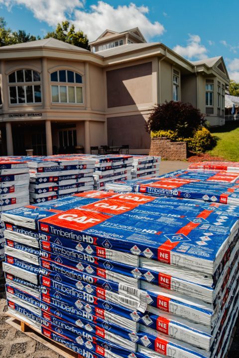 Stacks of blue and red roofing shingles in front of a light brown building with windows.