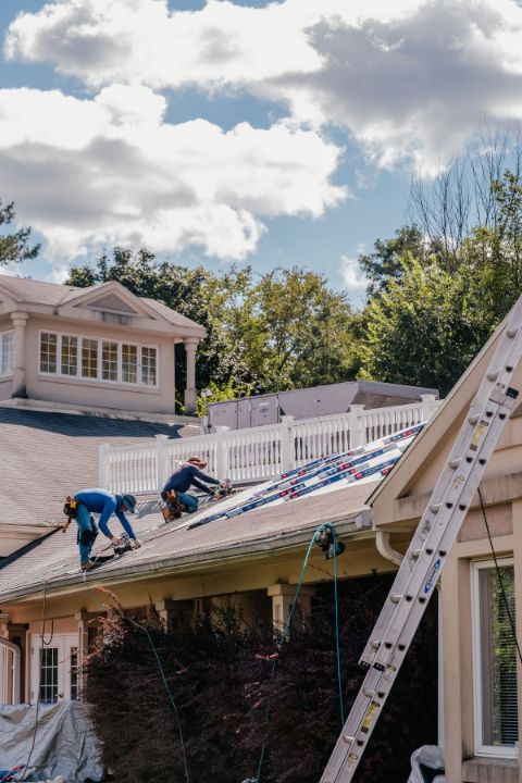 Two workers installing solar panels on a house roof. Blue uniforms, ladder visible. Overcast, daylight.