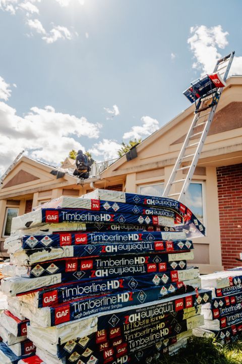 Roofing materials stacked on the ground. A ladder leads to a roof where a worker is. Sunny day.
