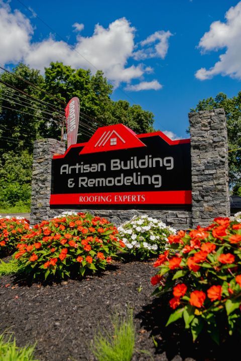 Sign for Artisan Building & Remodeling with red and black text, surrounded by stone pillars, flowers, and blue sky.
