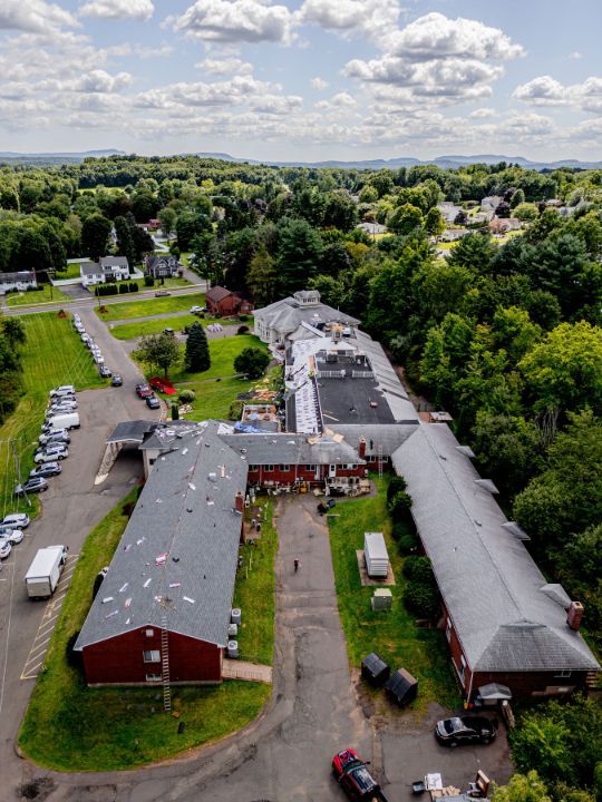 Aerial view of a long, red-roofed building, possibly a nursing home, surrounded by green trees and a parking lot.