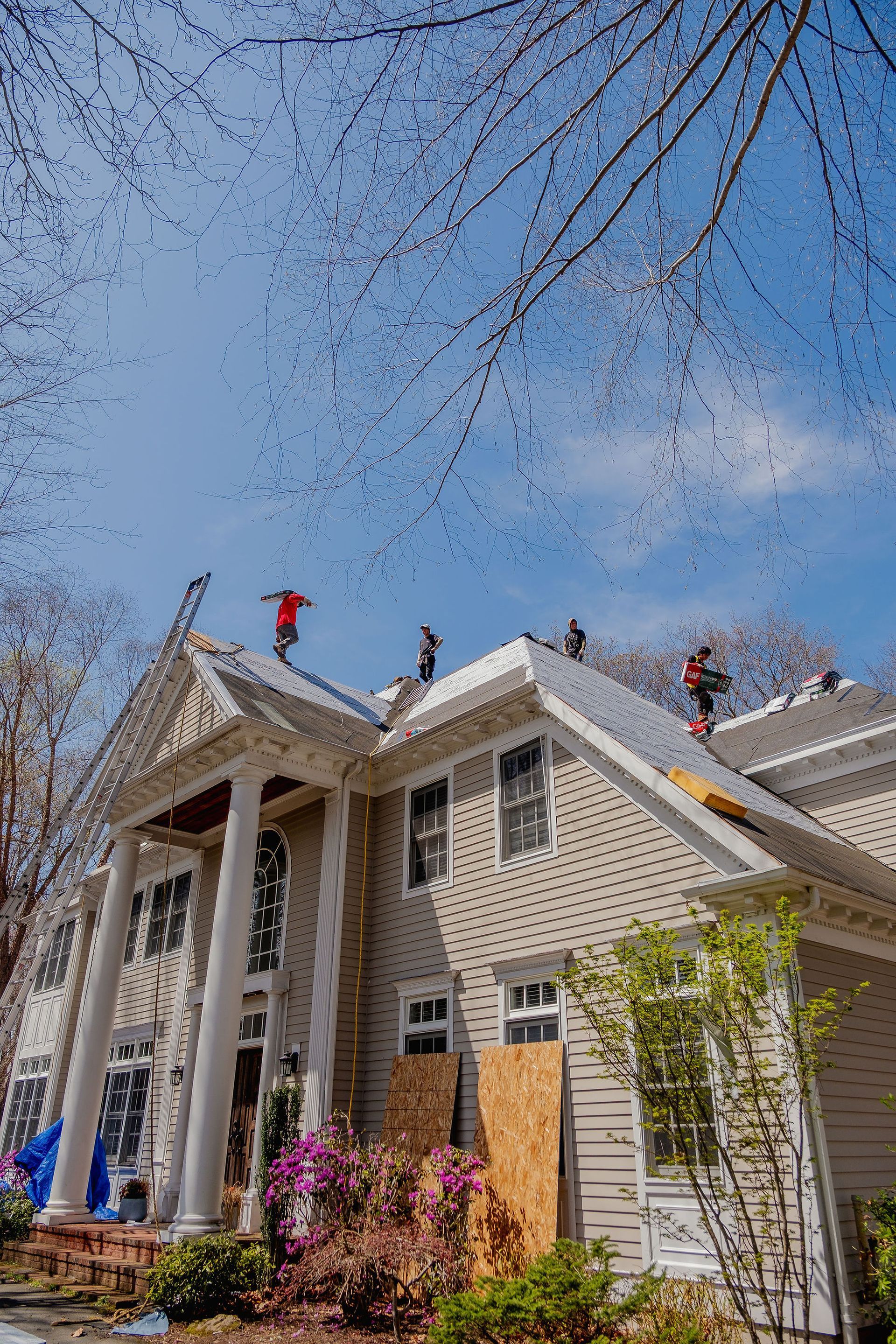 Roofers work on a two-story home with columns and a partially covered roof. Spring blooms below.