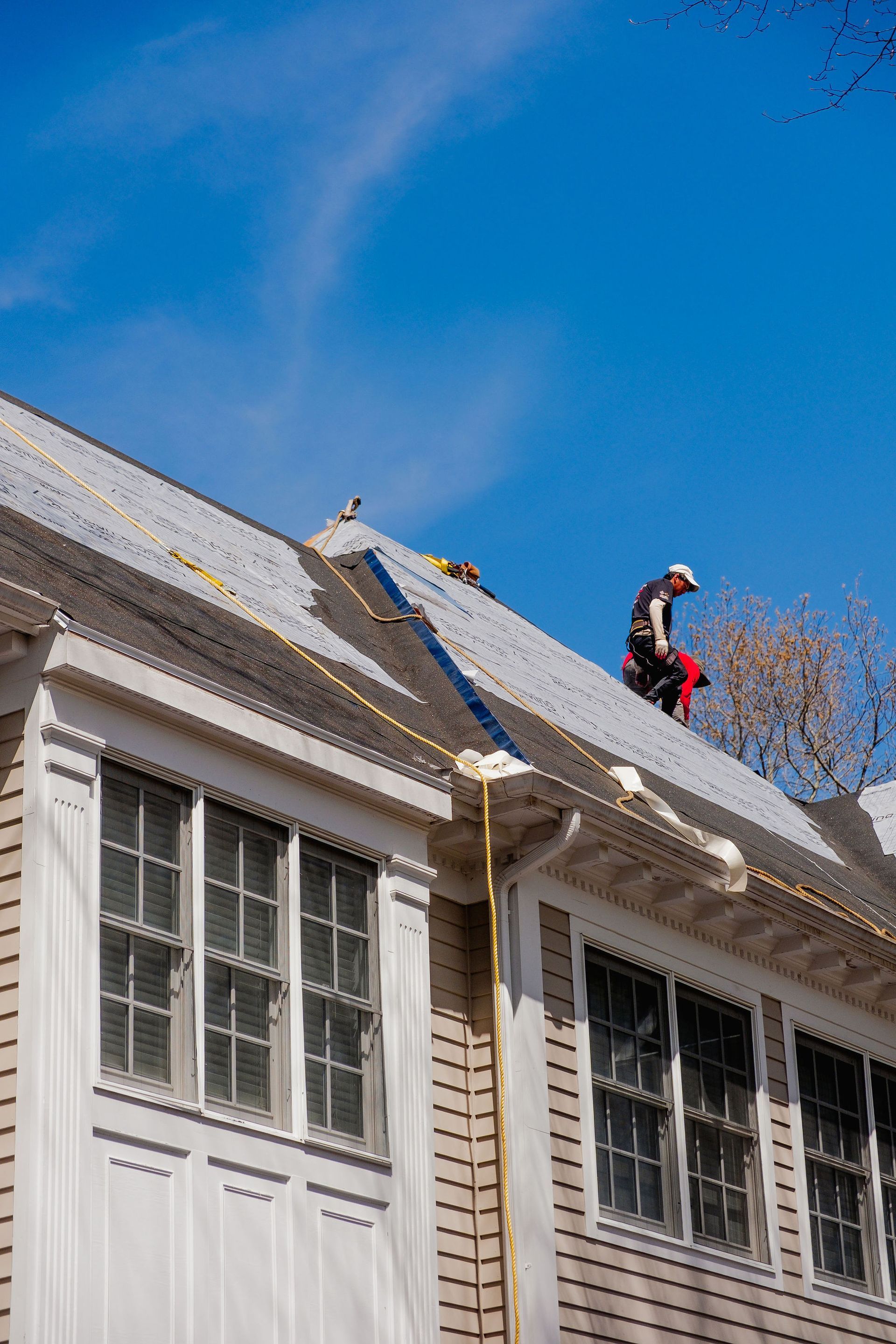 Person working on a roof, blue sky background. The house has white trim and multiple windows.